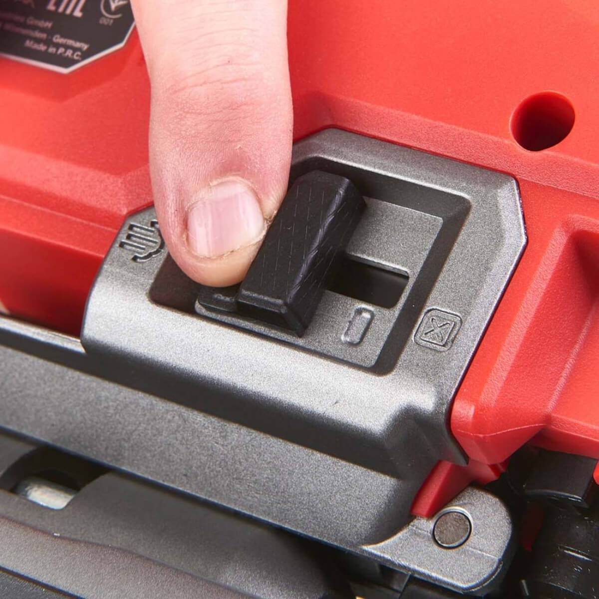 Close-up of a person pressing a switch on a red and grey and Red Milwaukee Jigsaw held by a hand on a white background.